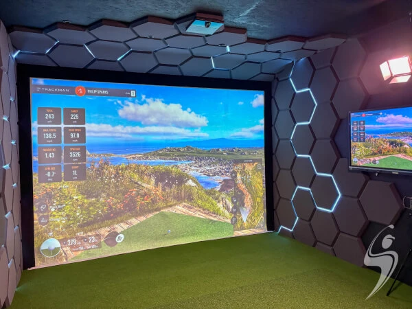 Golf simulator room with a large screen displaying a scenic golf course, hexagonal wall panels, and a smaller screen on the side.