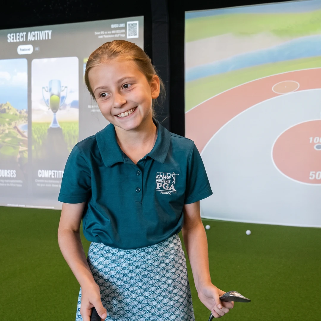 Young girl smiling while holding a golf club indoors, standing on green turf with interactive golf screens in the background.