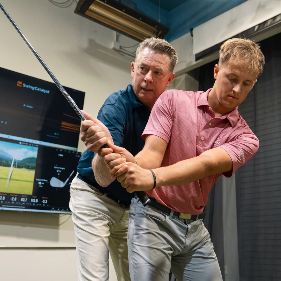 A golf instructor guides a young golfer's swing indoors, with a monitor displaying analysis data in the background.
