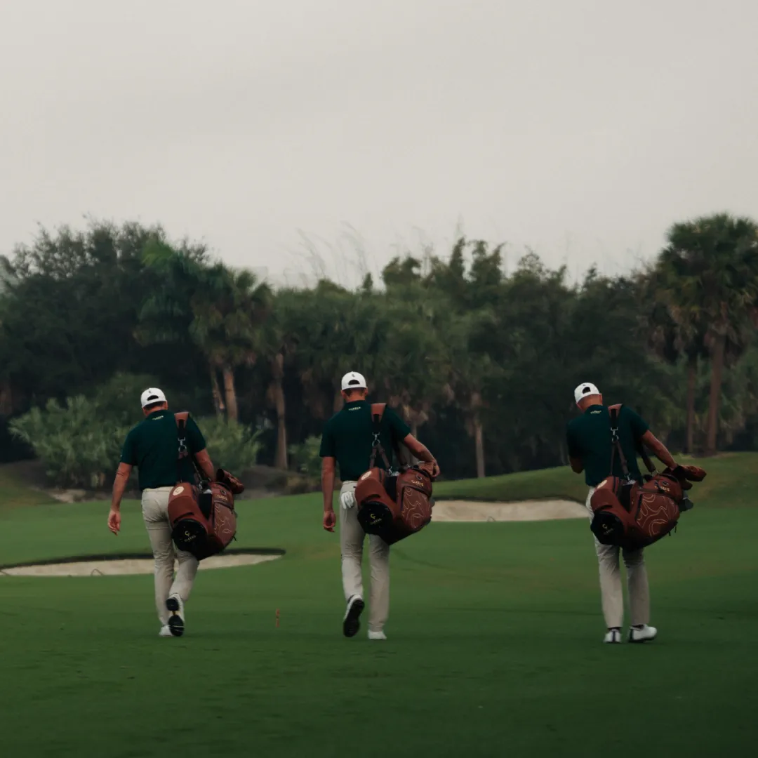 Three golfers in green shirts and white caps walk on a golf course, each carrying a bag under a cloudy sky.