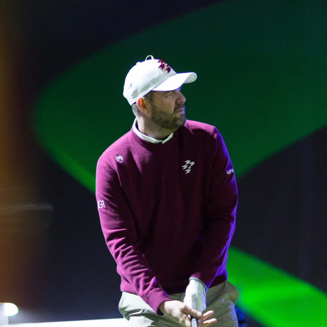 Golfer in a maroon sweater and white cap prepares for a shot on a dark, softly lit course with a green backdrop.
