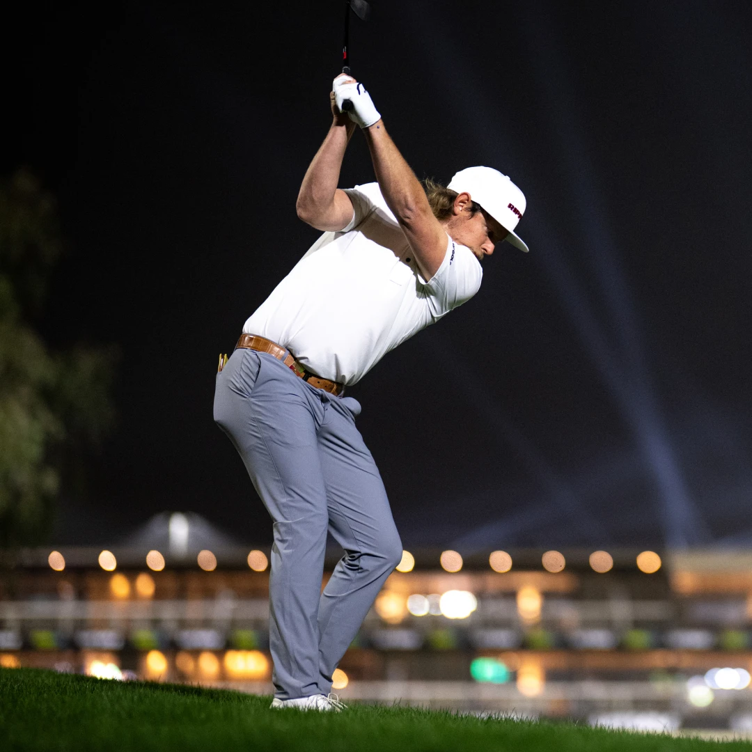 Golfer in mid-swing wearing a white shirt and cap, standing on grass with bright stadium lights in the background at night.