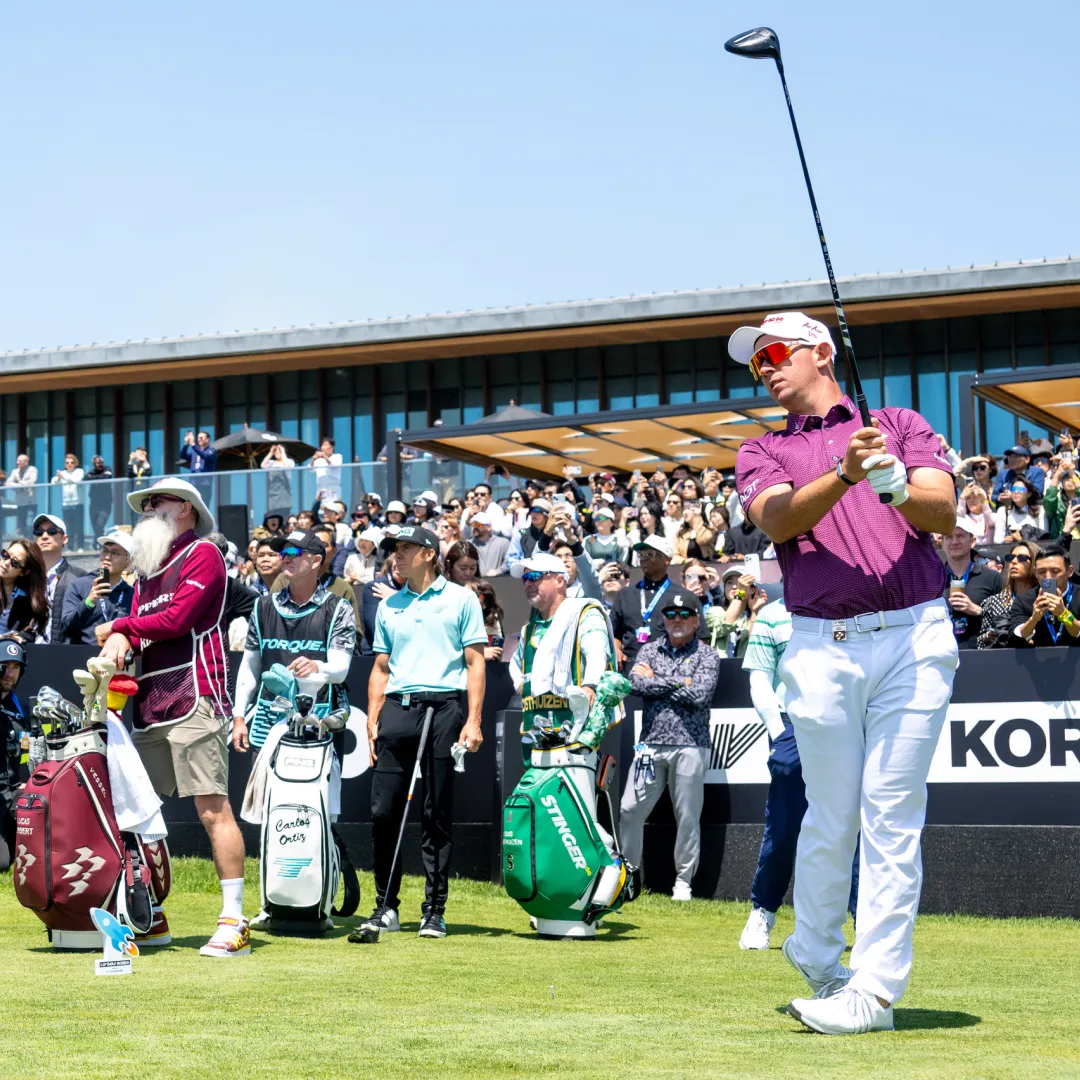 Golfer in a purple shirt watches his shot at a tournament, surrounded by caddies and spectators. Golf bags are visible on the grass.