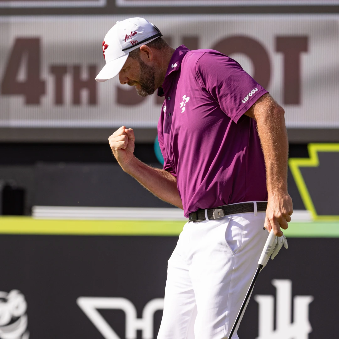 Golfer in a purple shirt and white pants celebrates with a fist pump, holding a putter on a golf course.