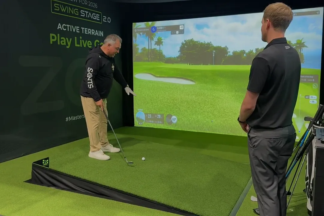 Two people in a golf simulator setup. One prepares to swing, while the other looks on. Virtual green and screen visible.