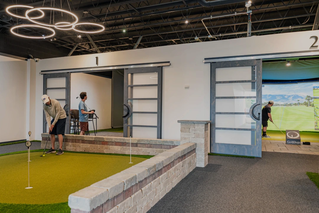 Indoor golf facility with two individuals practicing; one on a putting green, another using a golf simulator. Modern lighting and sliding doors.