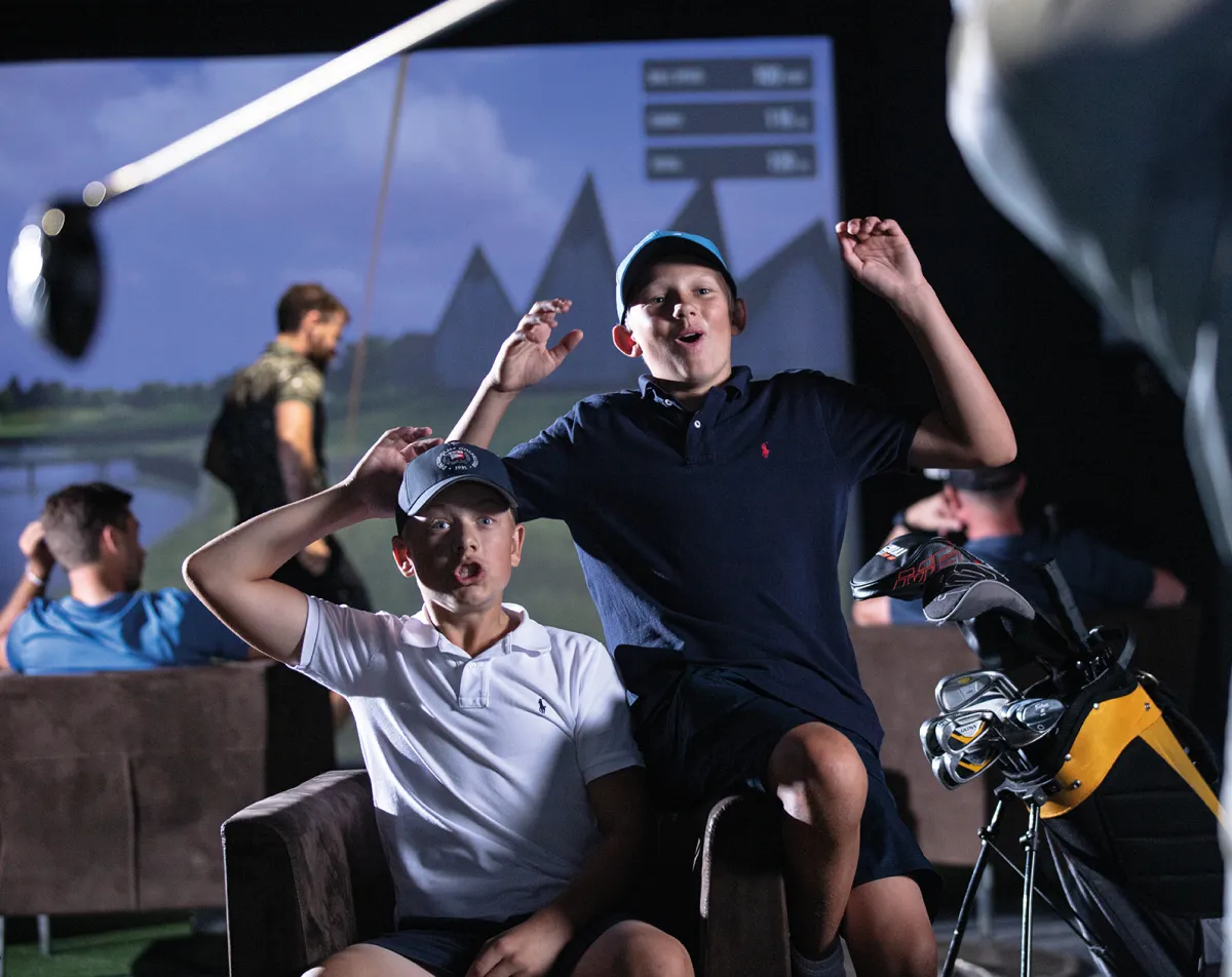 Two boys cheer enthusiastically in a virtual golf simulation room, with golf clubs nearby and a projected golf course in the background.