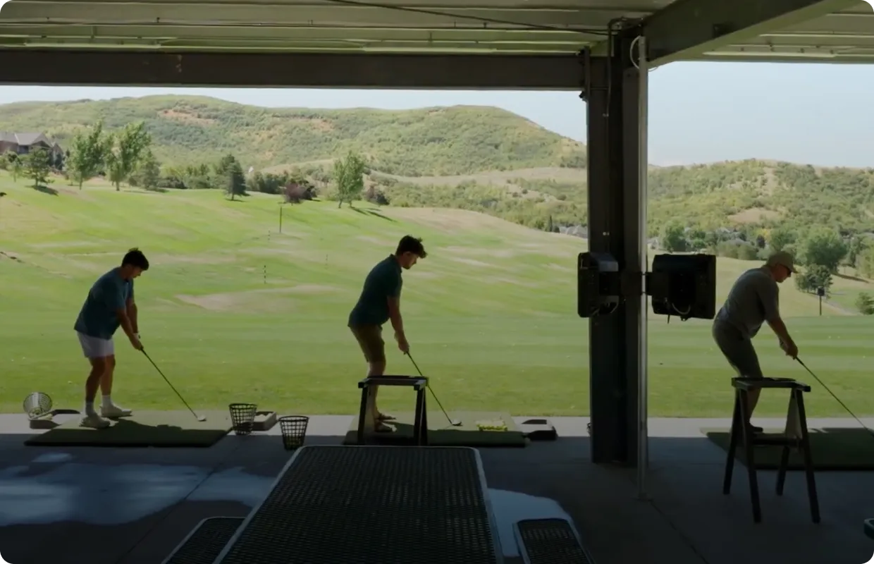 Three people practice golf swings at an indoor driving range with a view of a lush, rolling green landscape and hills in the background.
