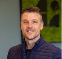 Smiling man with short hair wearing a dark quilted jacket, standing indoors with a green and black abstract artwork in the background.