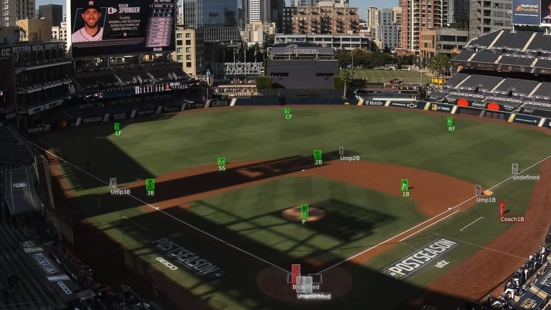 Aerial view of a baseball field with labeled positions and a large screen displaying player information, set in an urban backdrop.