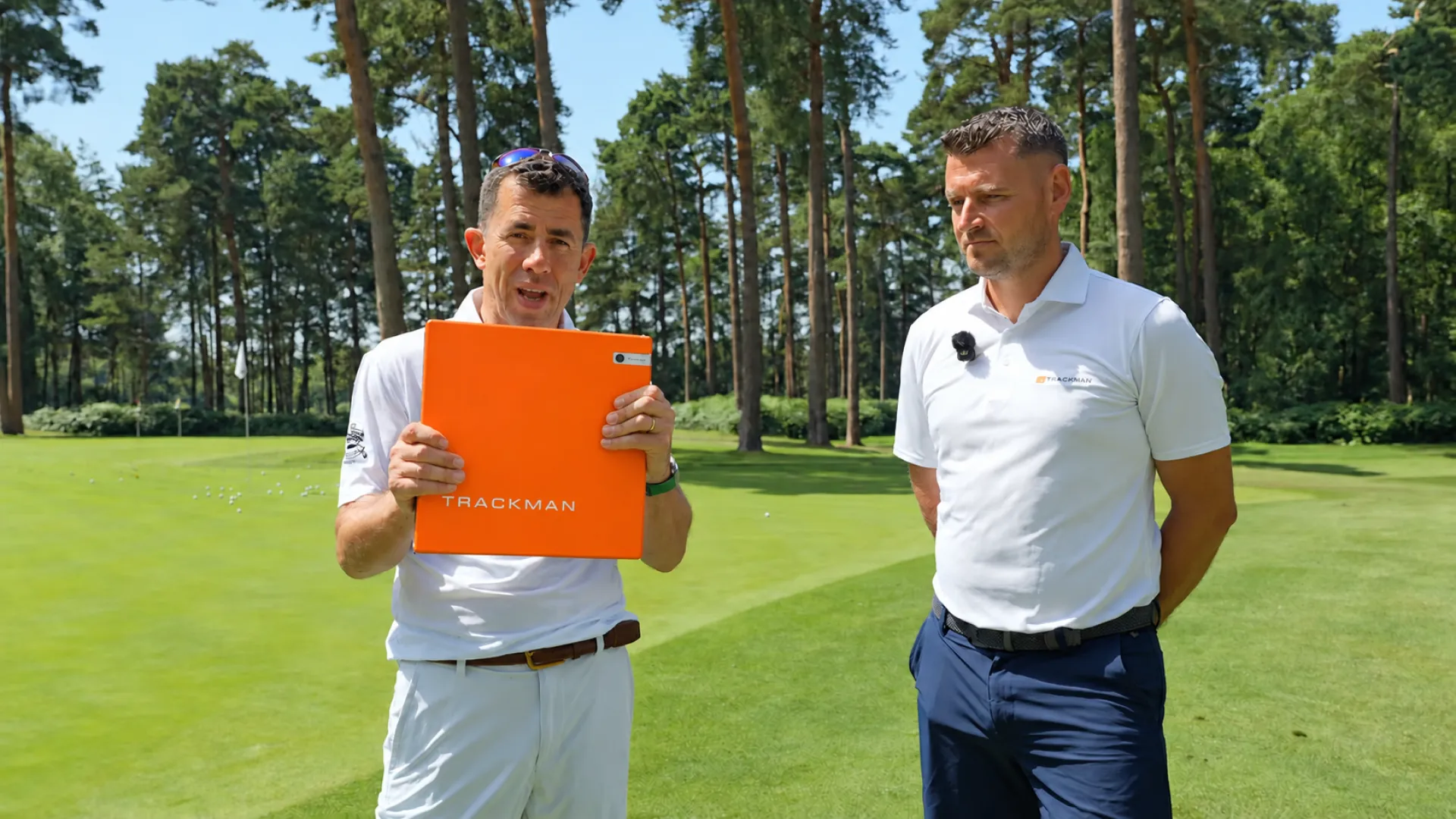 Two men stand on a golf course; one holds an orange Trackman device. Both wear white shirts, and trees are visible in the background.
