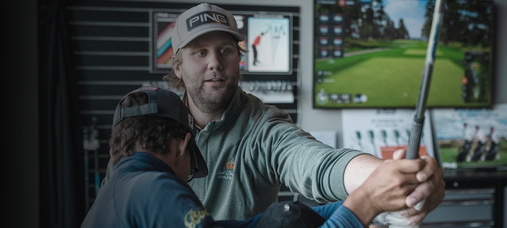 A golf instructor guides a student indoors, showing a swing technique. Both wear hats, and golf equipment is visible in the background.