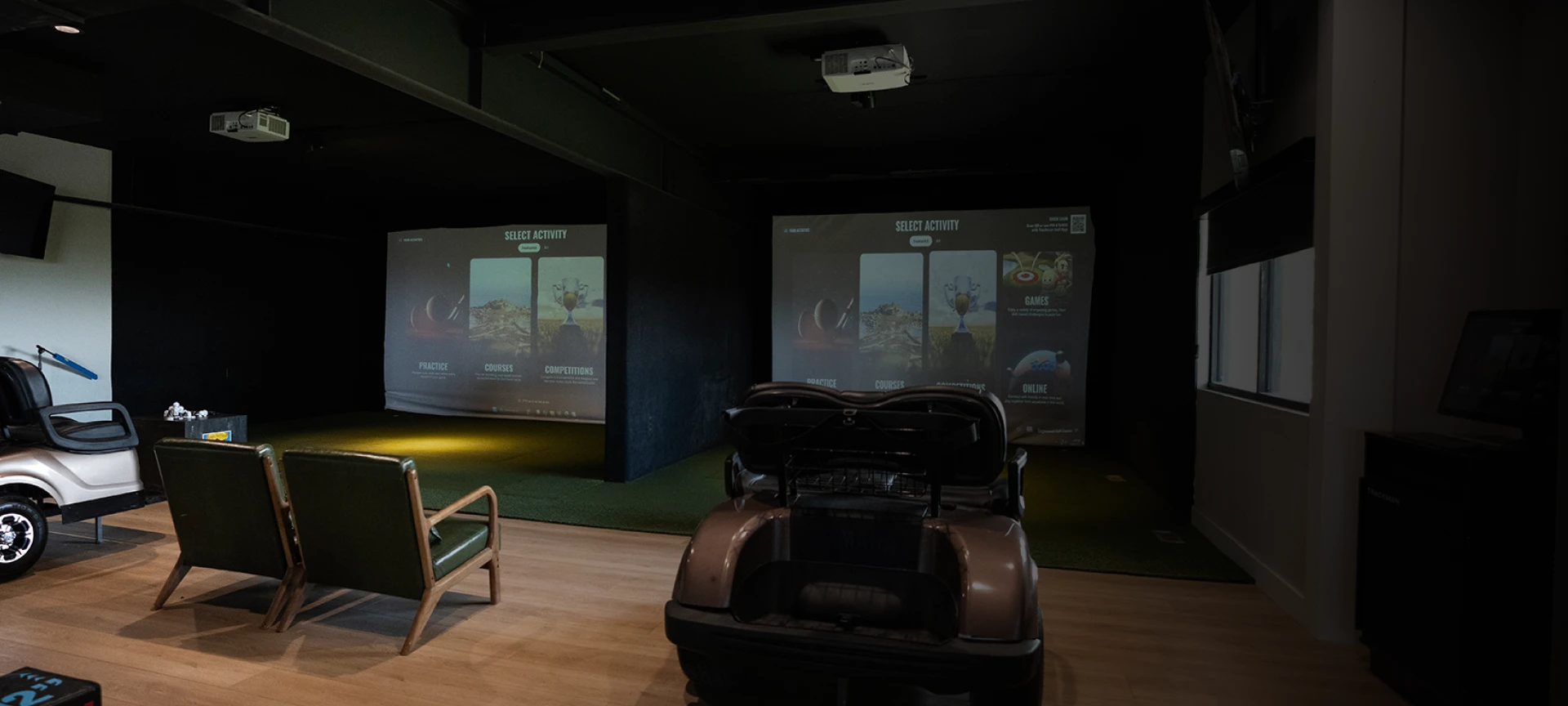 Indoor golf simulator setup with two screens displaying golf courses, two green chairs, and a golf cart in a dimly lit room.