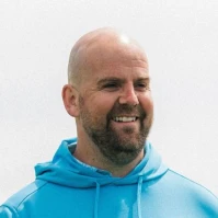 Tom Davies with a beard smiles while wearing a light blue hoodie against a plain white background.