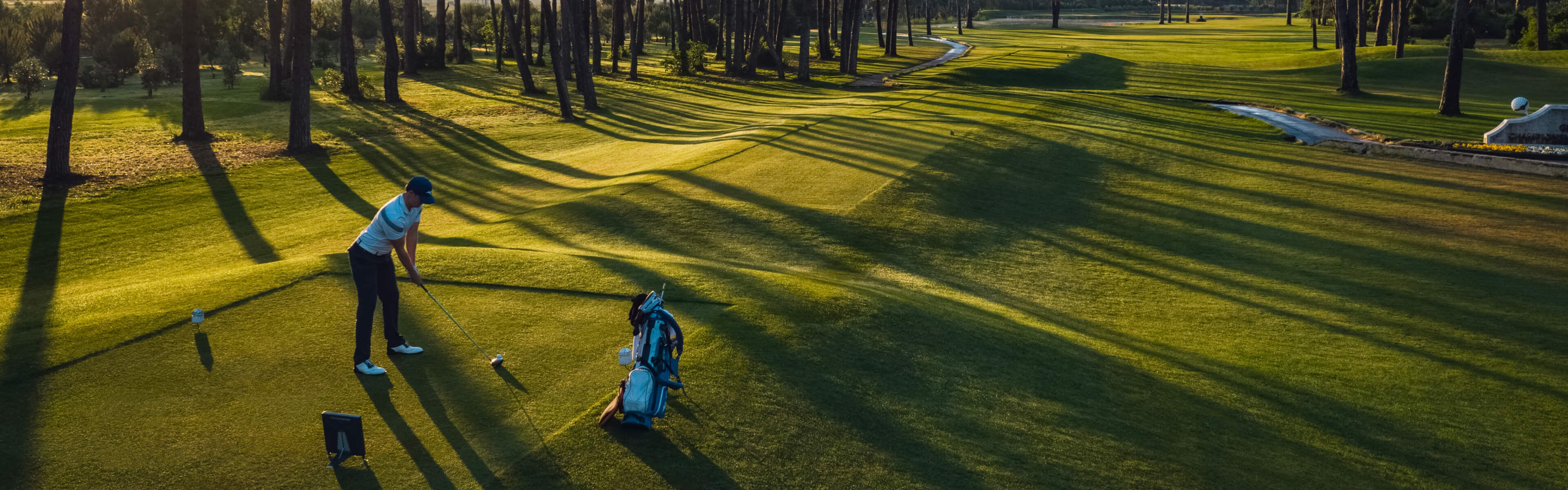 Trackman golfer preparing to swing on a lush, sunlit golf course with long tree shadows and a clear sky in the background.