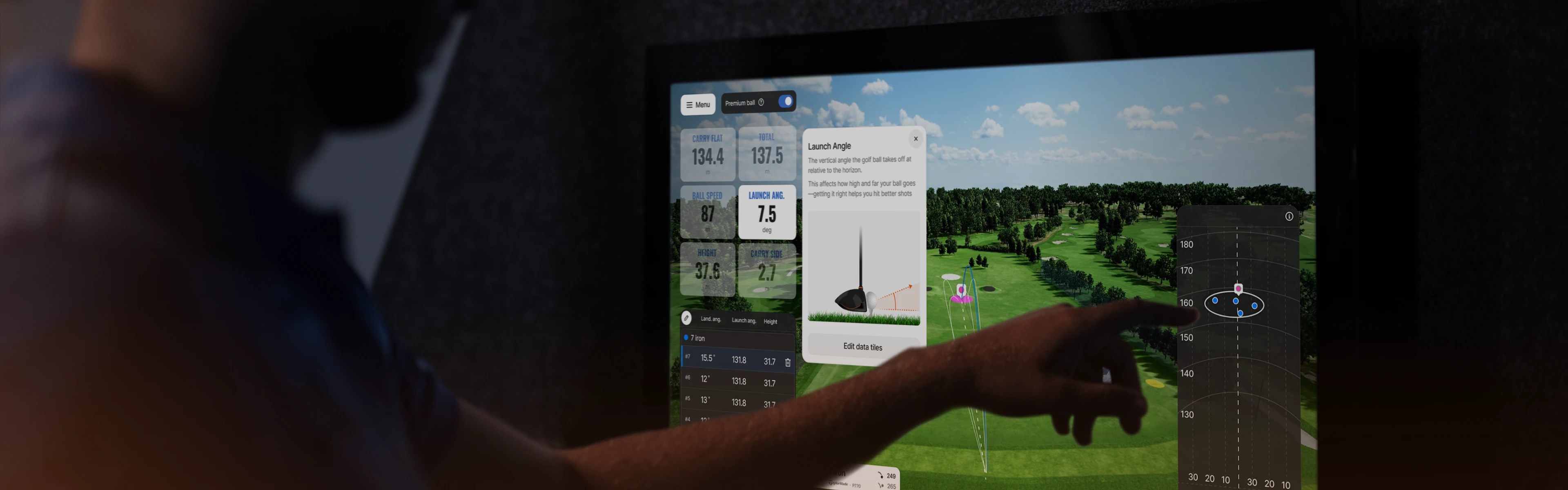 Person pointing at a digital golf simulator screen displaying launch angle and ball trajectory data against a virtual golf course backdrop.