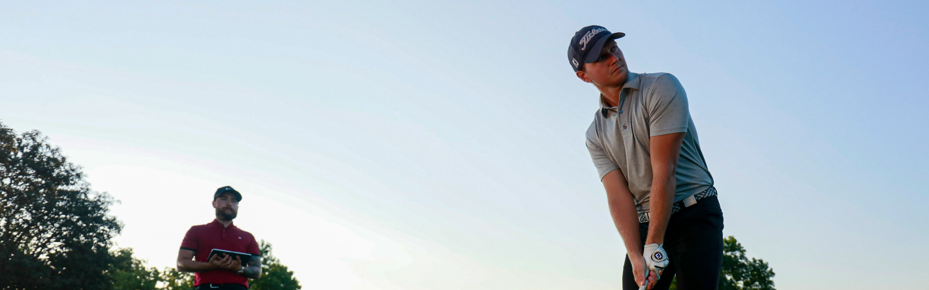 A golfer in a gray shirt and cap prepares to swing with a club, with a man in red holding a tablet in the background under a clear sky.