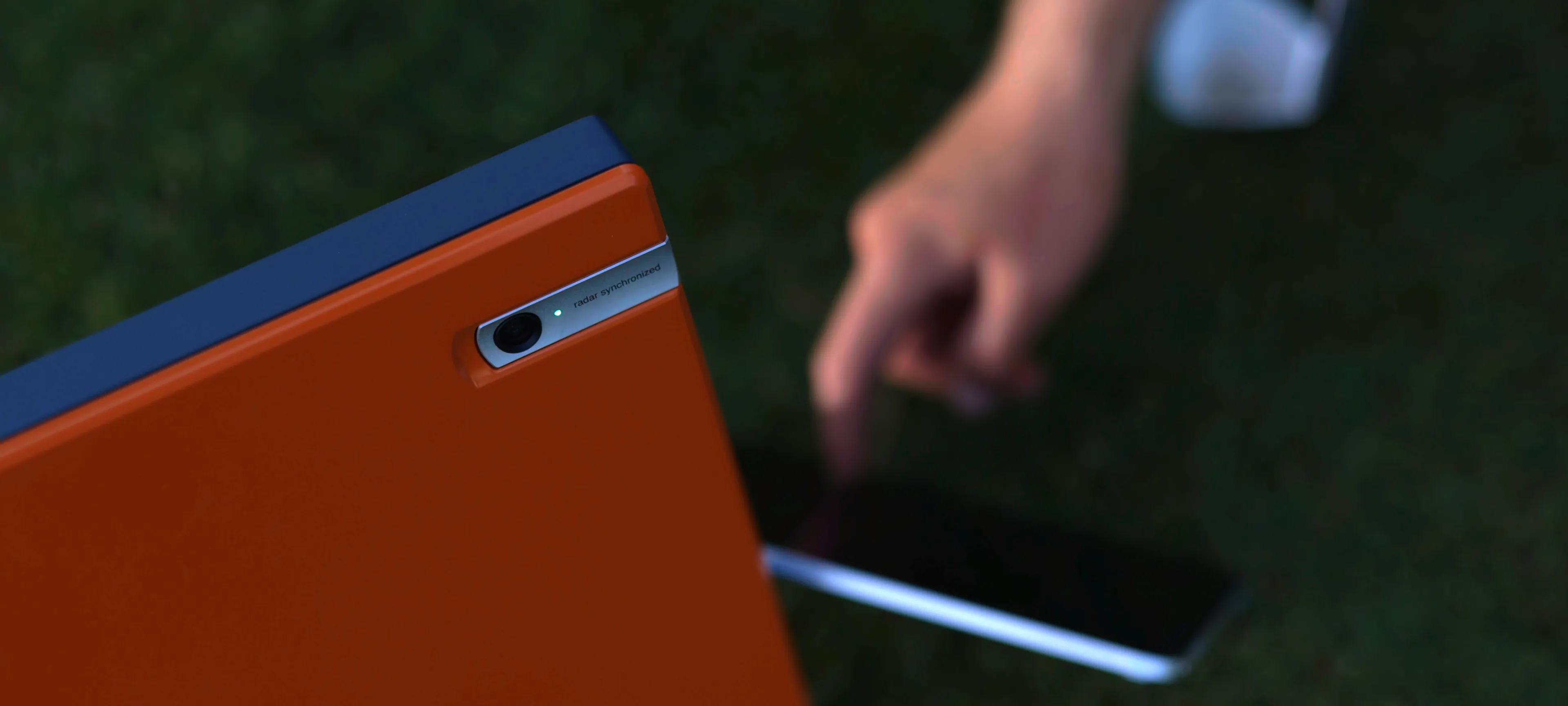 Close-up of a person interacting with an orange device with a camera and "radar synchronized" label, set against a blurred outdoor background.