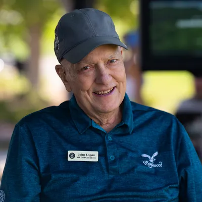 Elderly man in a blue polo and cap, smiling. He wears a name tag and is outdoors with blurred greenery in the background.