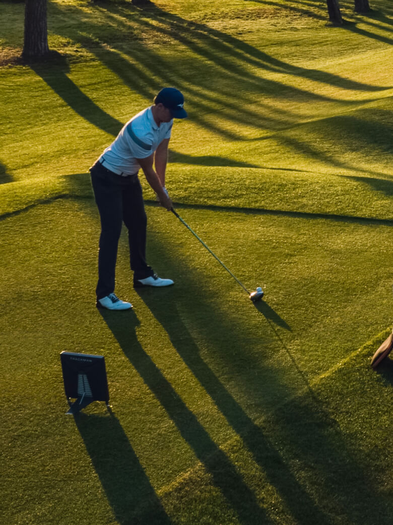 Trackman golfer in blue cap and white shirt preparing to swing on a sunlit golf course, with long shadows cast on the green grass.