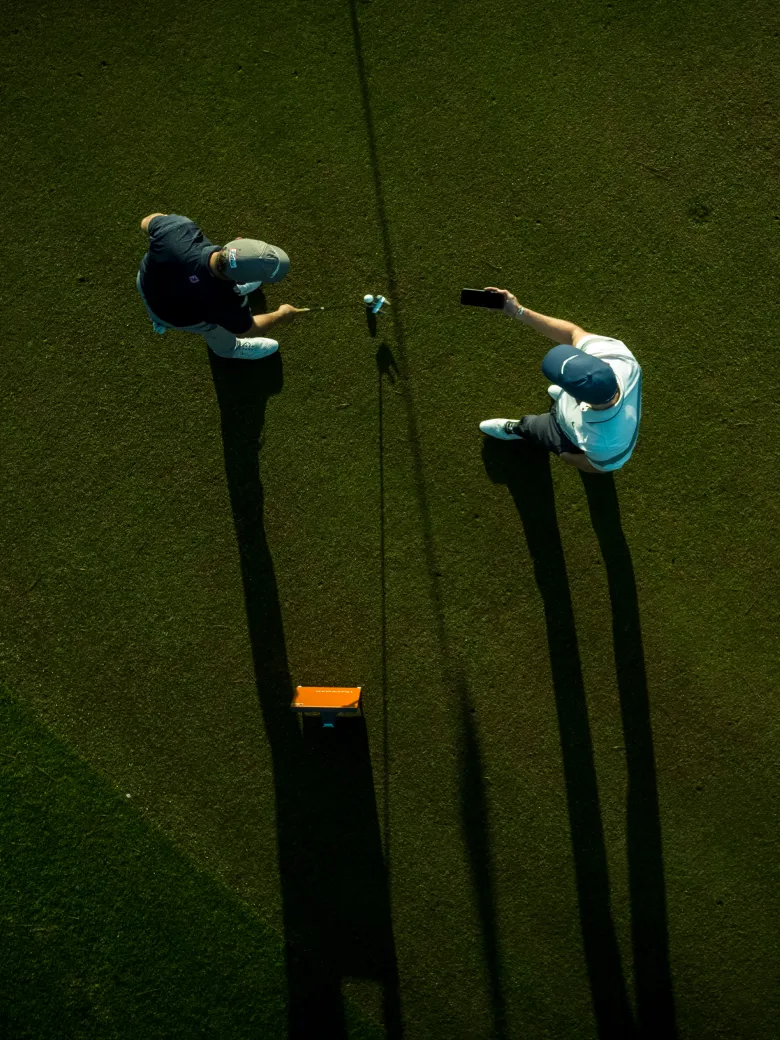Two golfers with caps on a green field captured from above, casting long shadows; one holds a club, the other a smartphone.