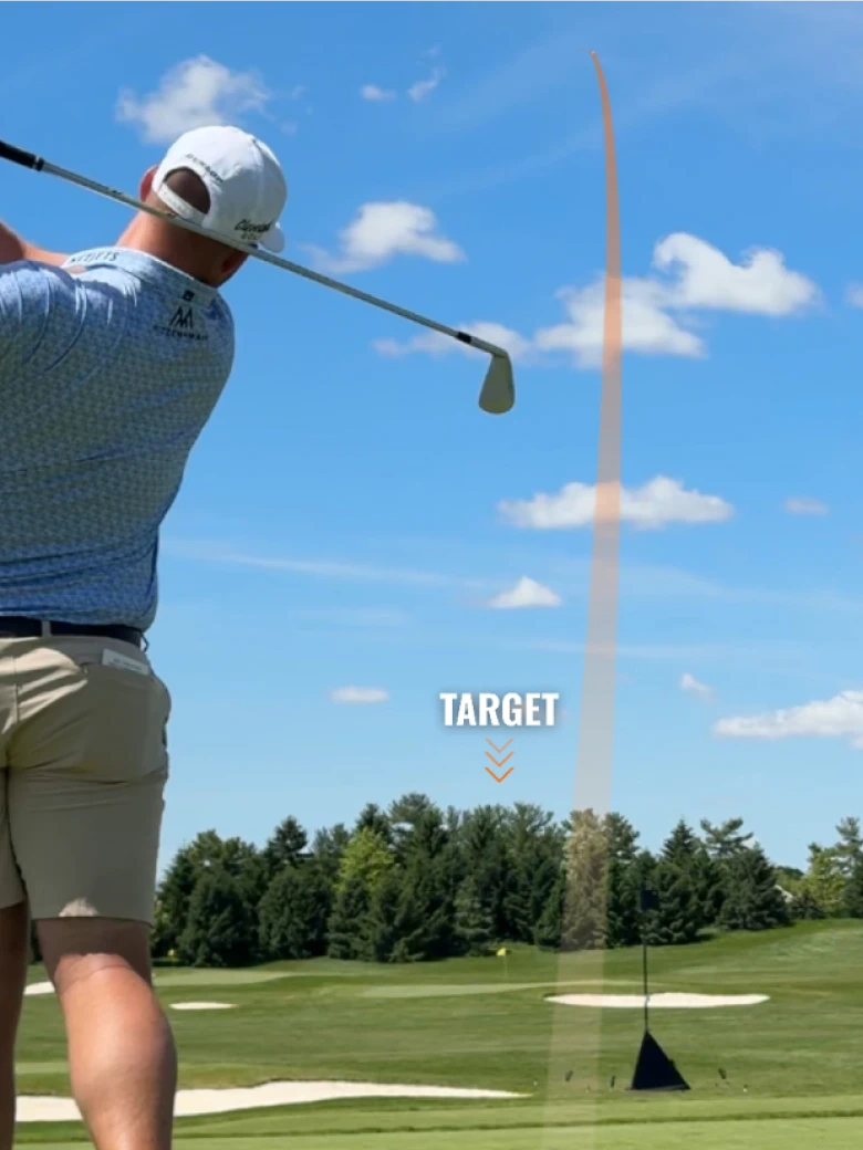 Golfer in a blue shirt and white cap swings at a golf course, with a trajectory line pointing at the target under a clear blue sky.