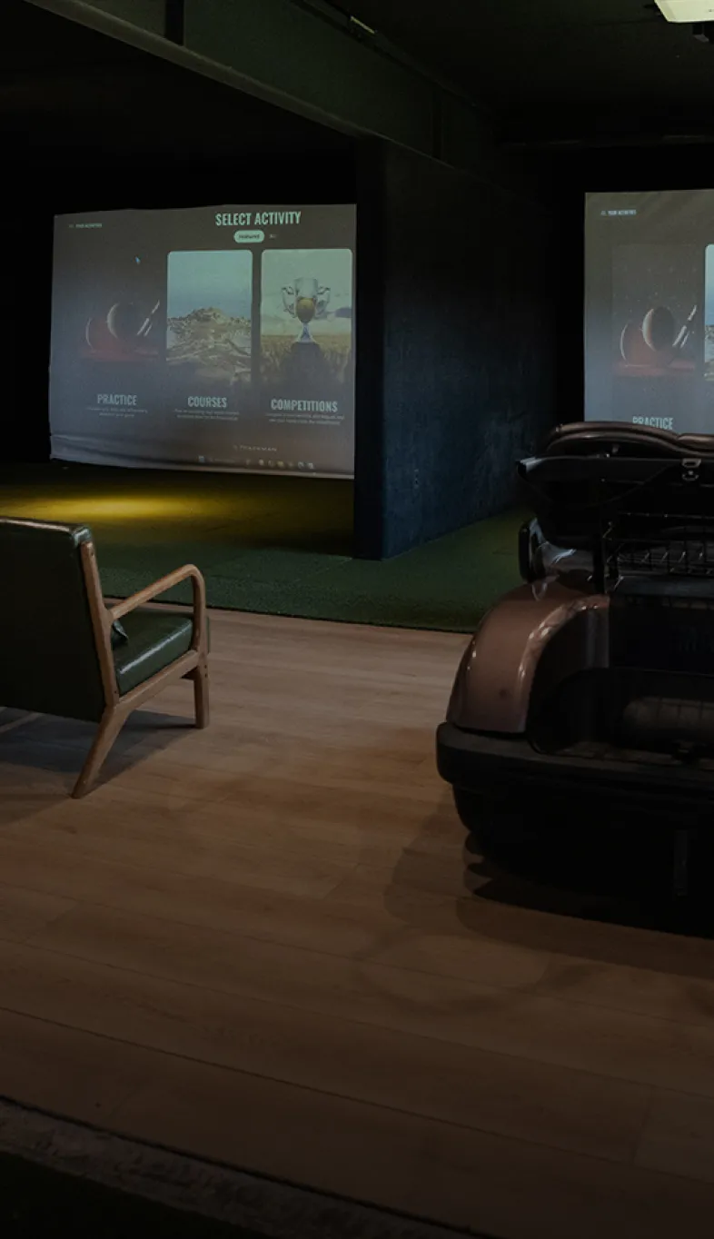Indoor golf simulator setup with two large screens displaying golf activities, a golf cart, and a wooden chair on a wooden floor.