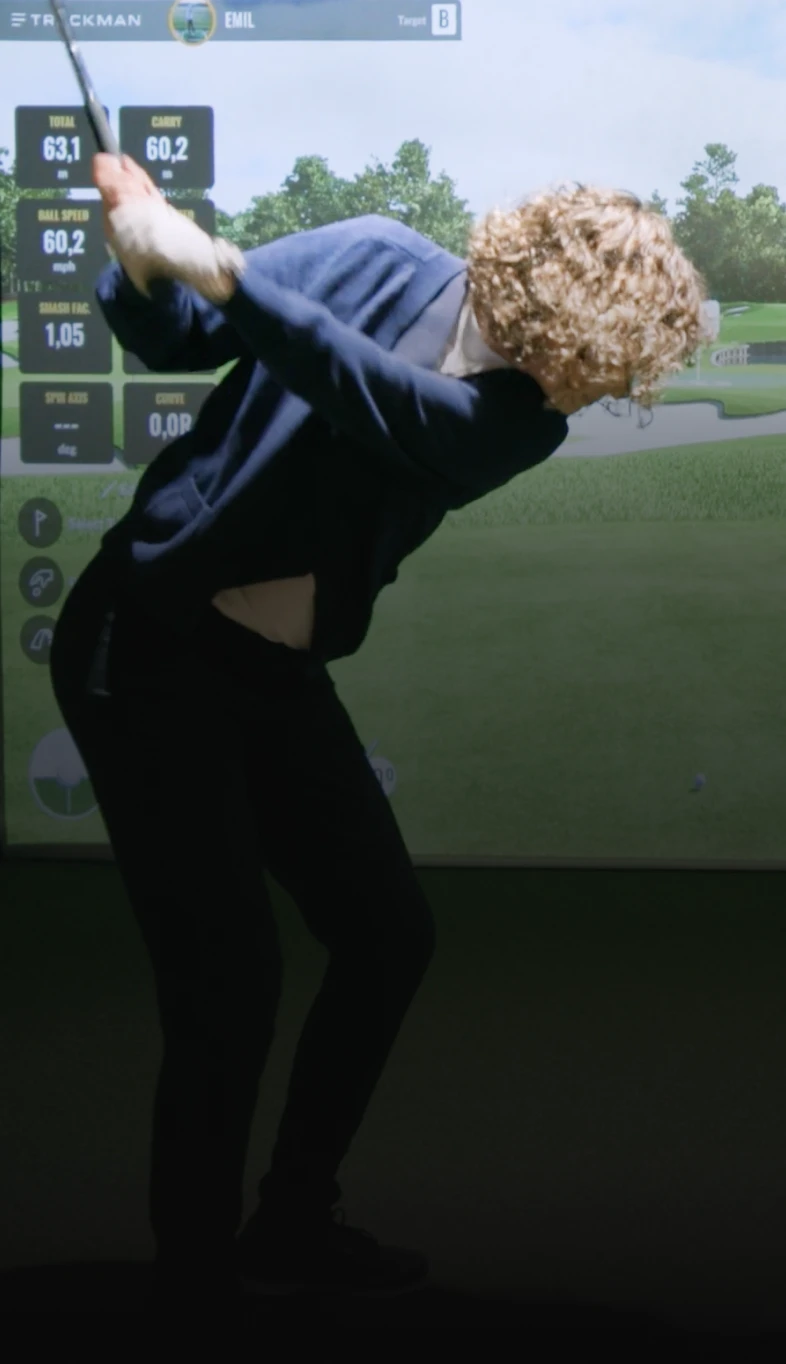 A person with curly hair swings a golf club indoors in front of a virtual golf simulator display.