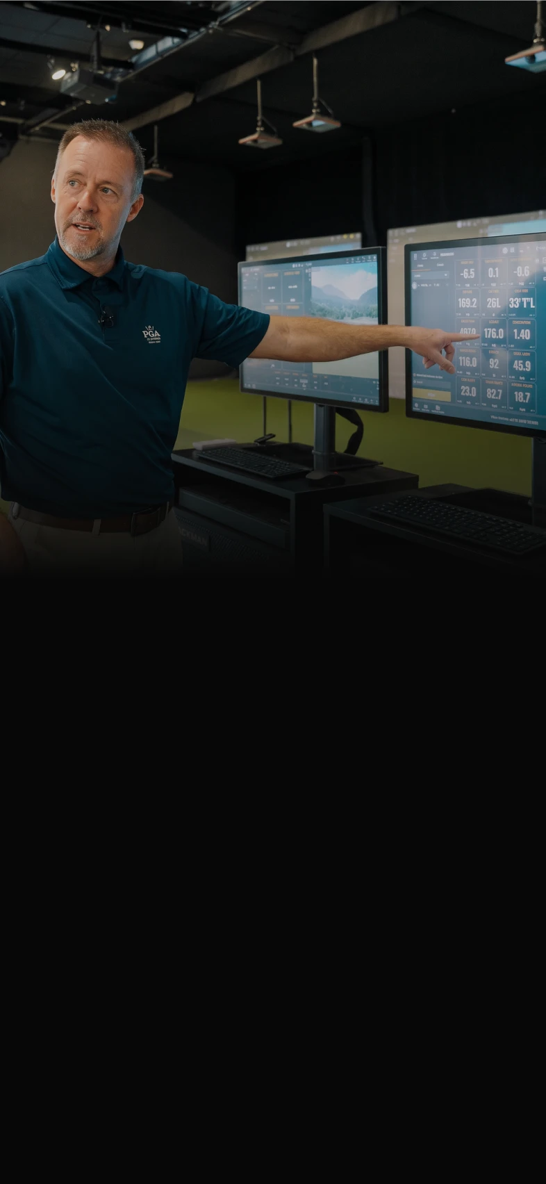 Man in a blue shirt pointing at data on a large monitor in a room with multiple screens and computers.