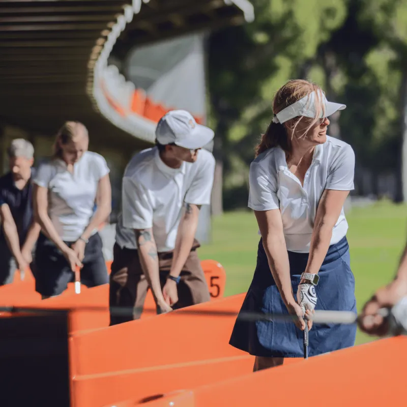 People in white shirts and visors practicing golf swings at a driving range, surrounded by trees in the background.