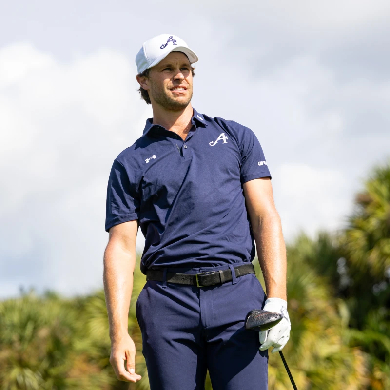 A man in a navy golf outfit and cap stands on a golf course, holding a club. Palm trees and a cloudy sky are in the background.