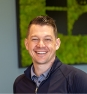 Smiling man in a navy sweater and blue shirt, standing indoors with a green abstract artwork in the background.