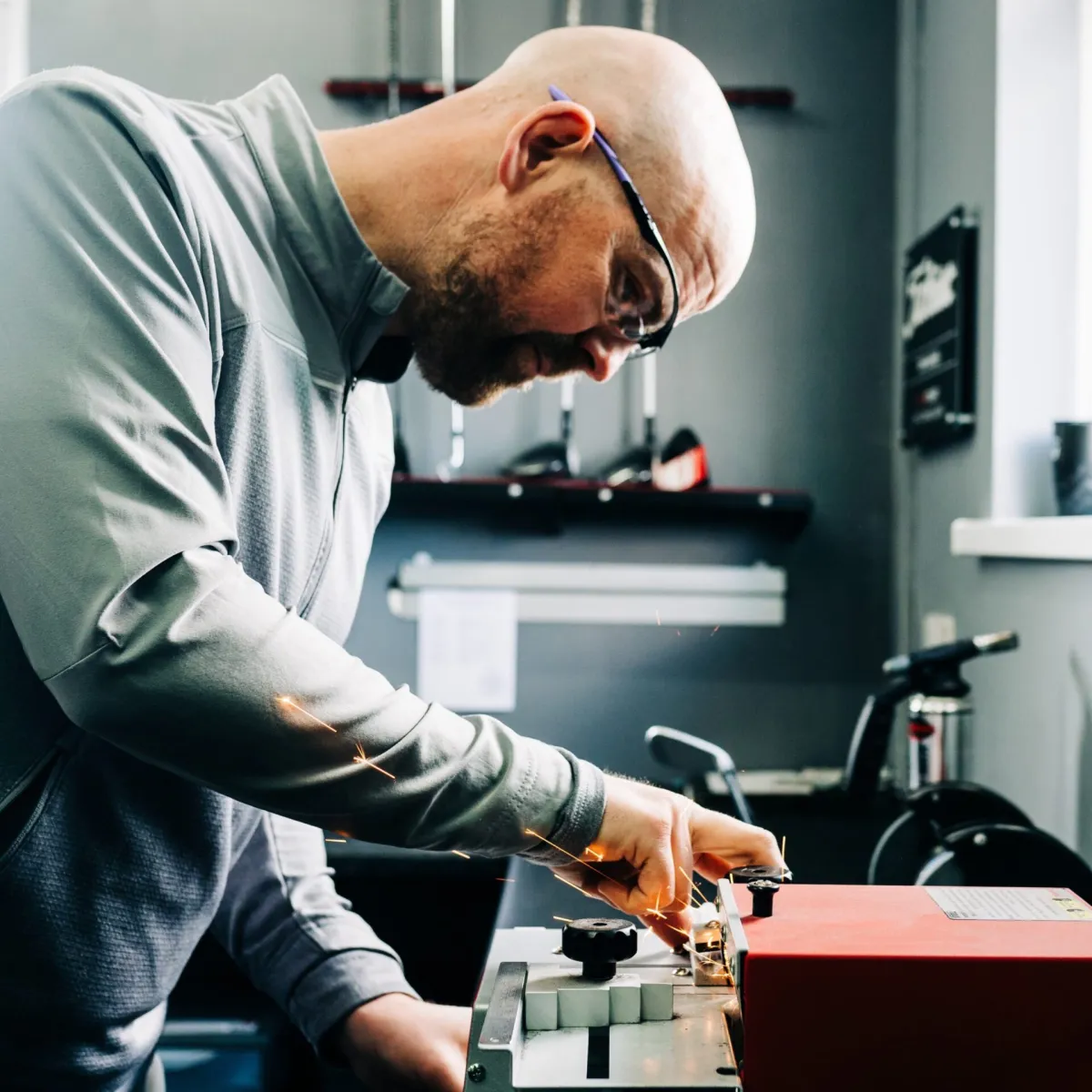 Man in safety glasses and gray jacket using a machine tool, producing sparks, in a workshop with shelves and tools in the background.