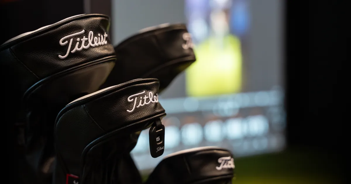 Close-up of several black golf club headcovers with "Titleist" branding, set against a blurred background displaying a screen.