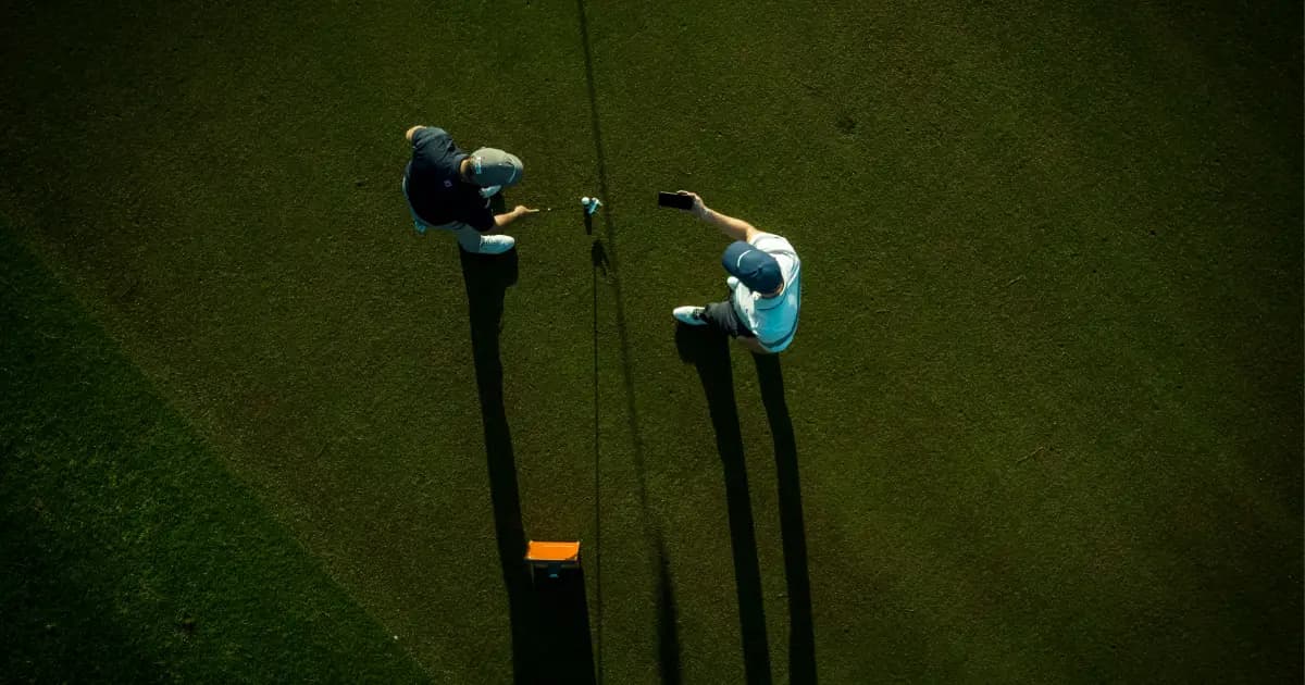 Overhead view of two golfers on a green, one taking a selfie. Long shadows and golf equipment visible.