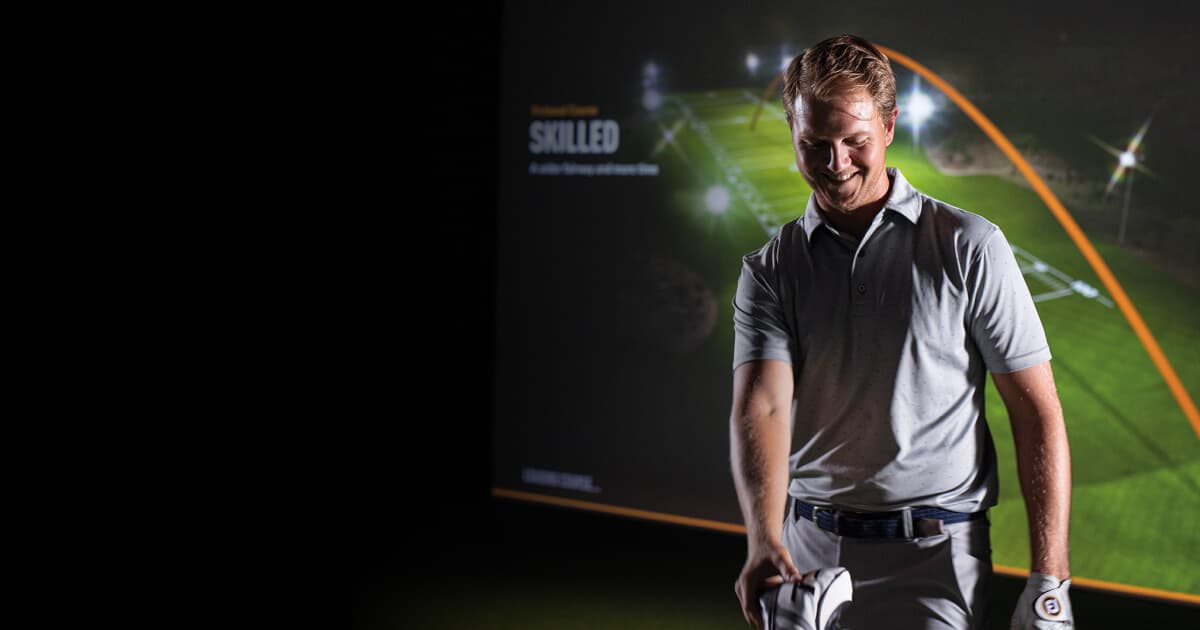 Smiling man in a golf Trackman simulator room, holding a club. Background shows a virtual golf course display.