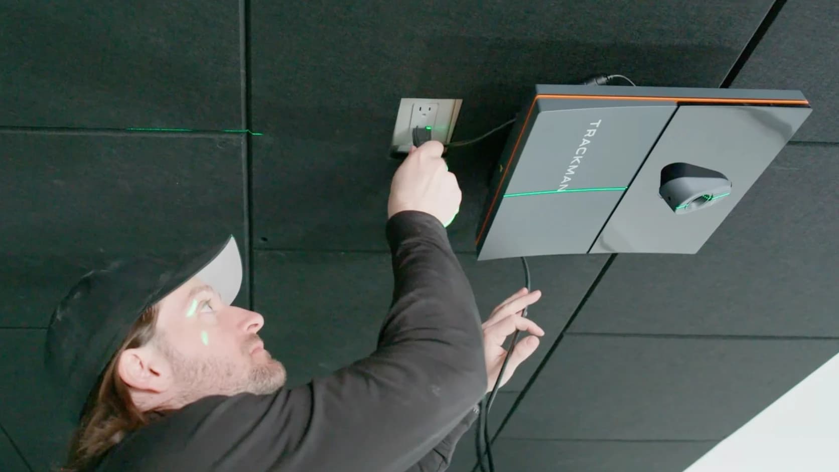Man wearing a cap installing a Trackman device on the ceiling, plugging it into a power outlet.