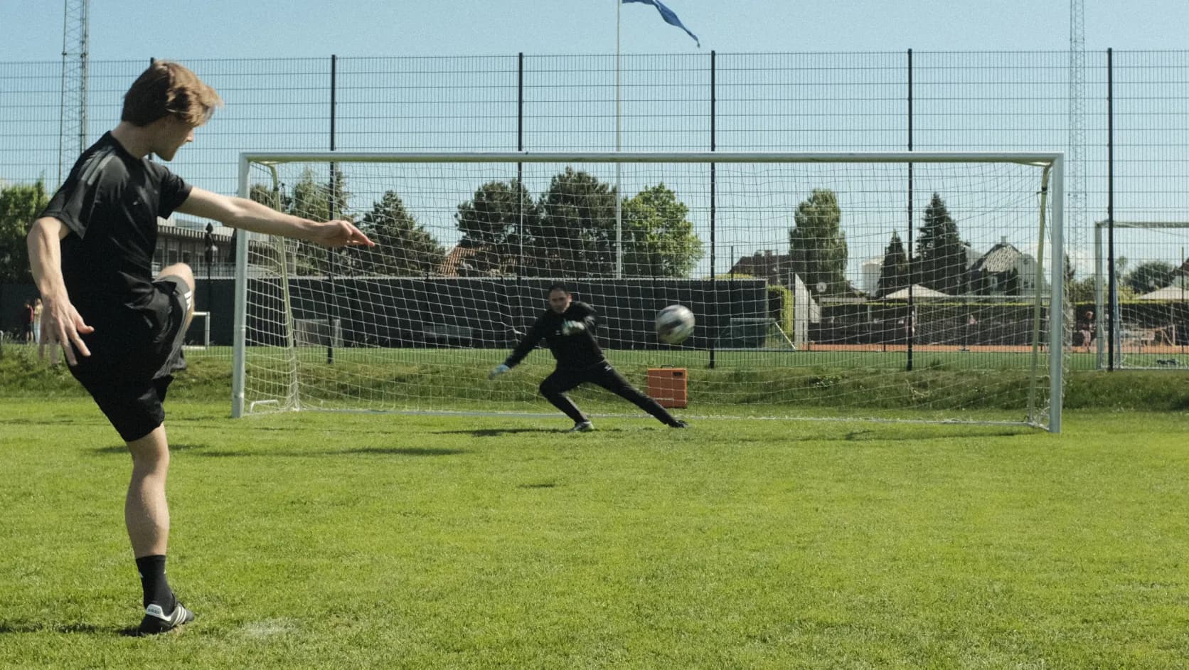 A soccer player kicks a ball towards a goalie attempting to save it, on a sunny day at a grass field with a netted fence background.