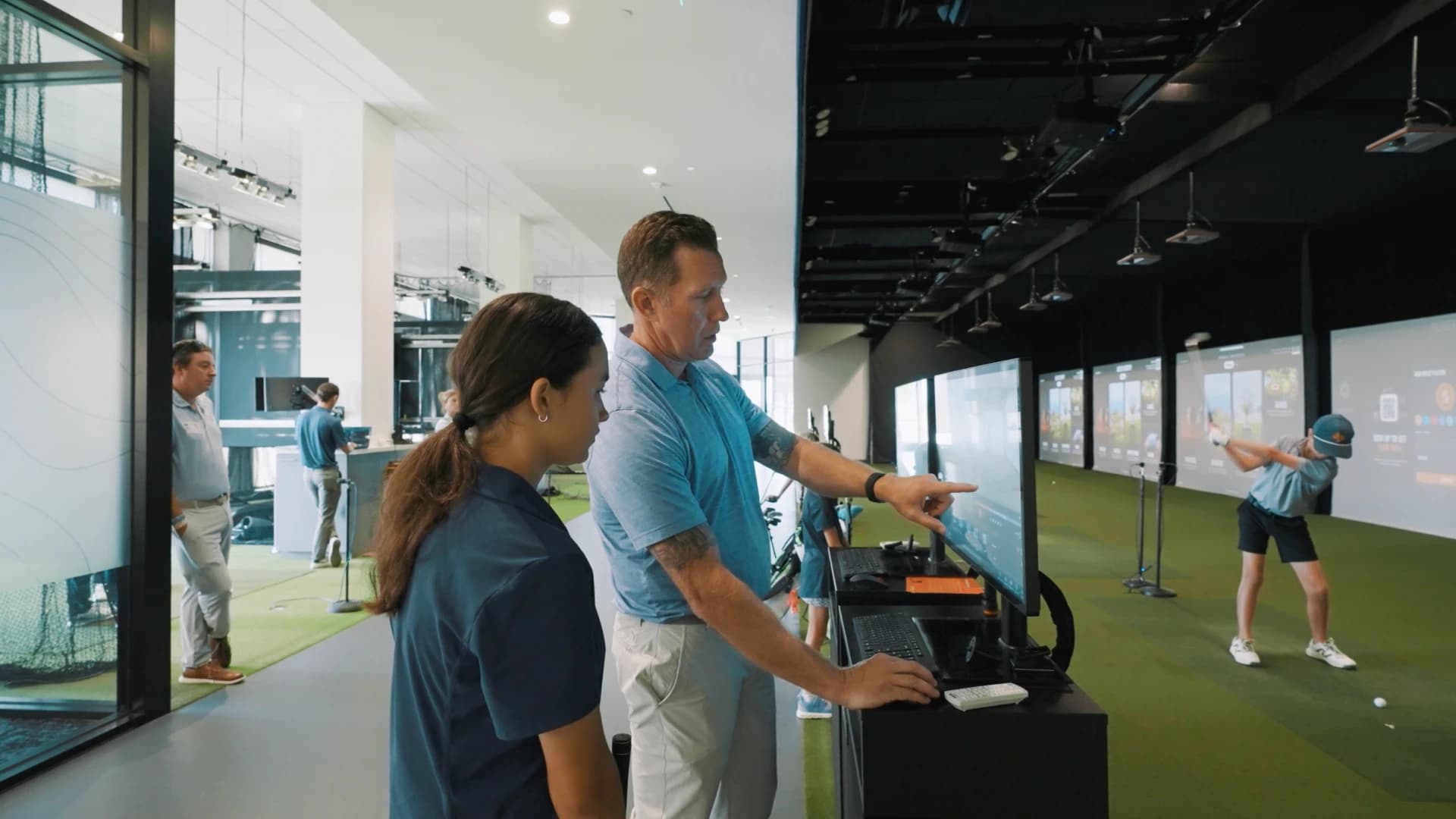 A man and a woman are looking at a monitor in a golf simulation facility, while a person swings a golf club in the background.