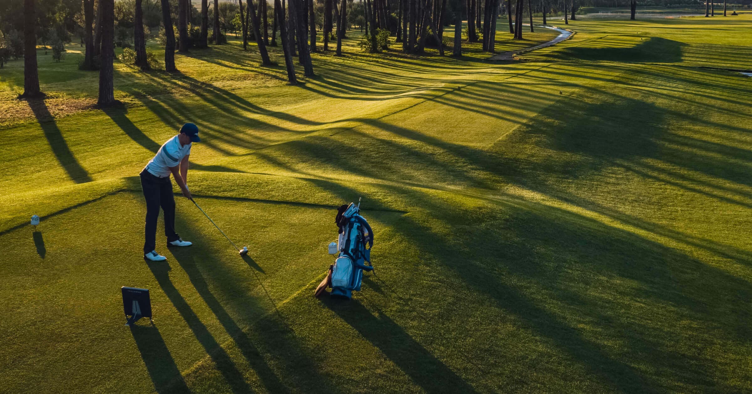 Golfer preparing to swing on a sunlit course, surrounded by trees casting long shadows on the grass.