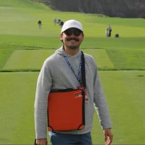 Brandon La Rosa, wearing a cap and sunglasses, stands on a golf course holding an orange case, with others in the background.