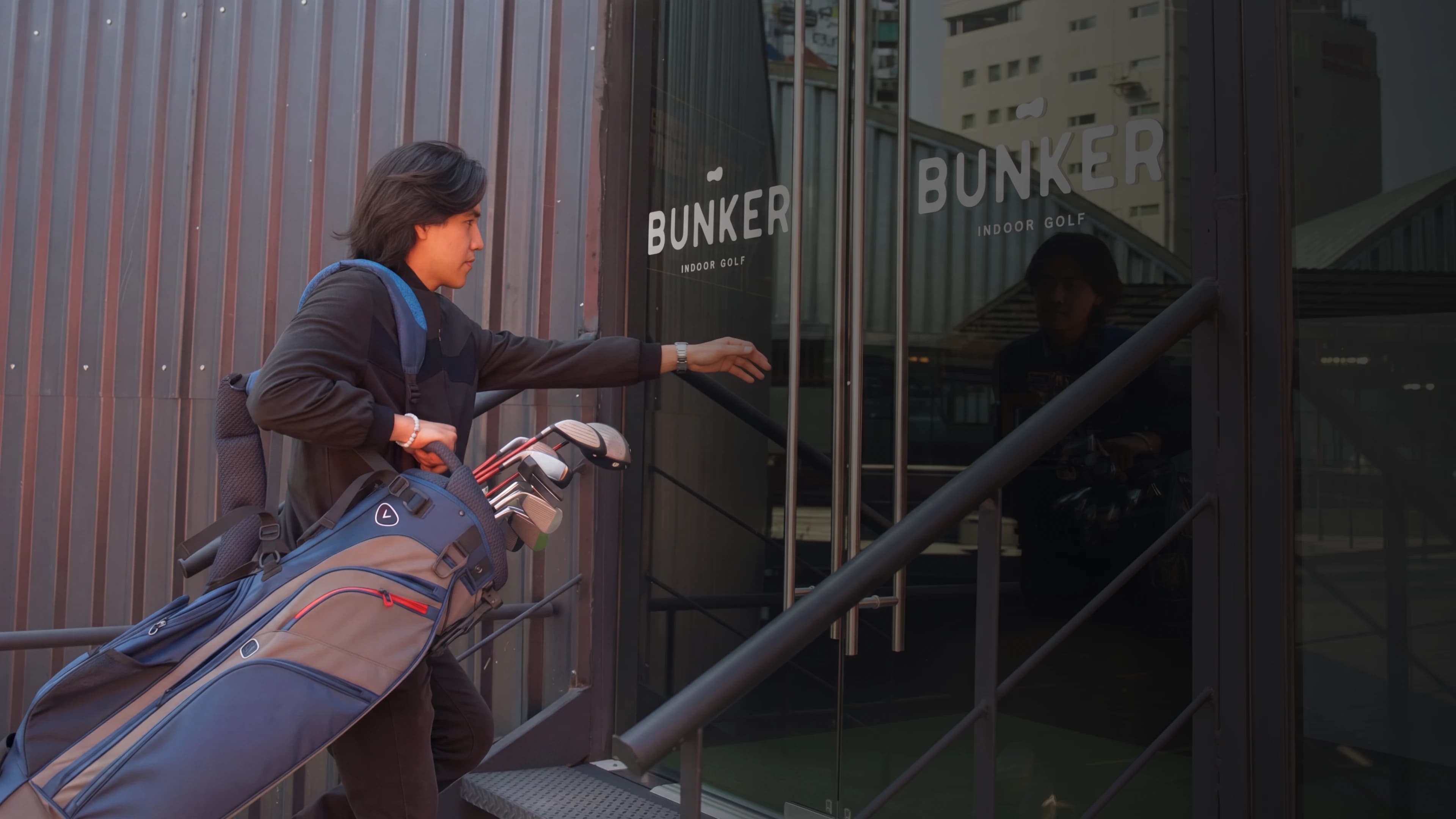 Person carrying golf clubs, reaching for the door of "Bunker Indoor Golf" in an urban setting.