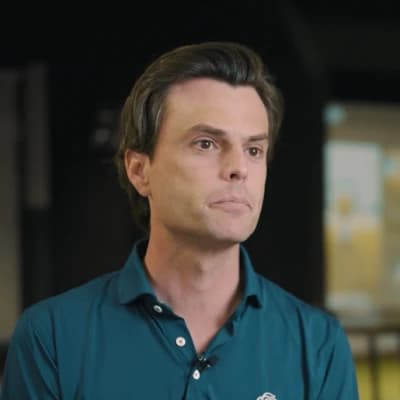 A man in a teal shirt stands in a dimly lit room, gazing forward with a neutral expression, blurred background behind him.