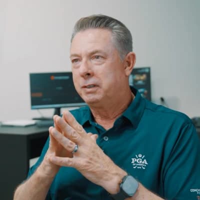 Man in a teal PGA polo shirt, seated with hands clasped, speaking in an office environment with computers in the background.