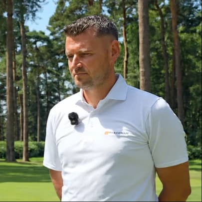 A man in a white polo shirt stands on a grassy area with tall trees in the background, looking off to the side.