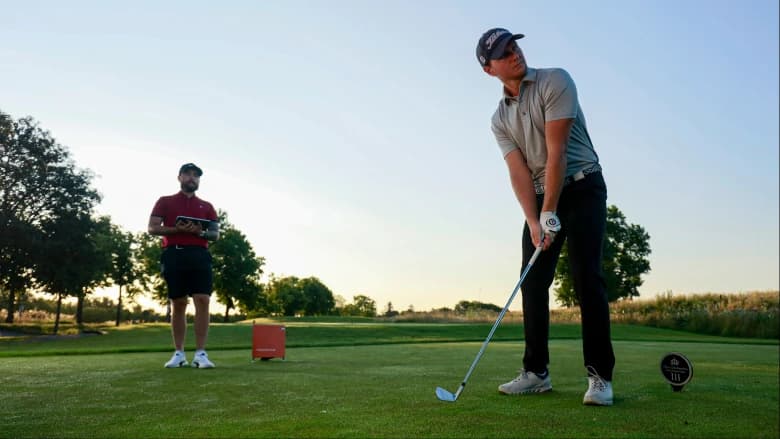 Golfer preparing to swing on a sunny course, with a person holding a tablet standing nearby under a clear blue sky.