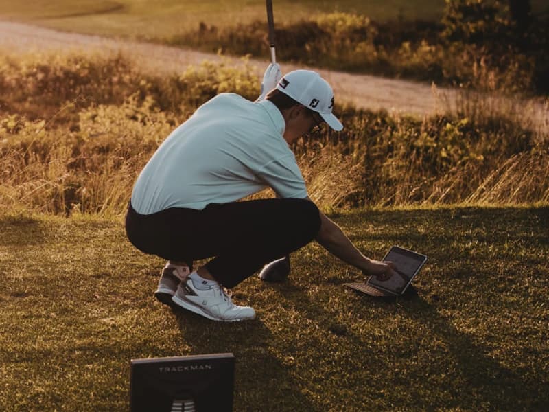 Golfer in white shirt and cap squatting on grass, using a laptop on a golf course at sunset, with a golf bag in the background.