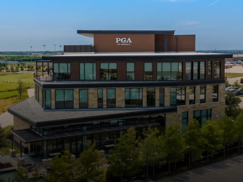 A modern multi-story building with large windows and "PGA of America" signage on top, surrounded by greenery and a parking lot.