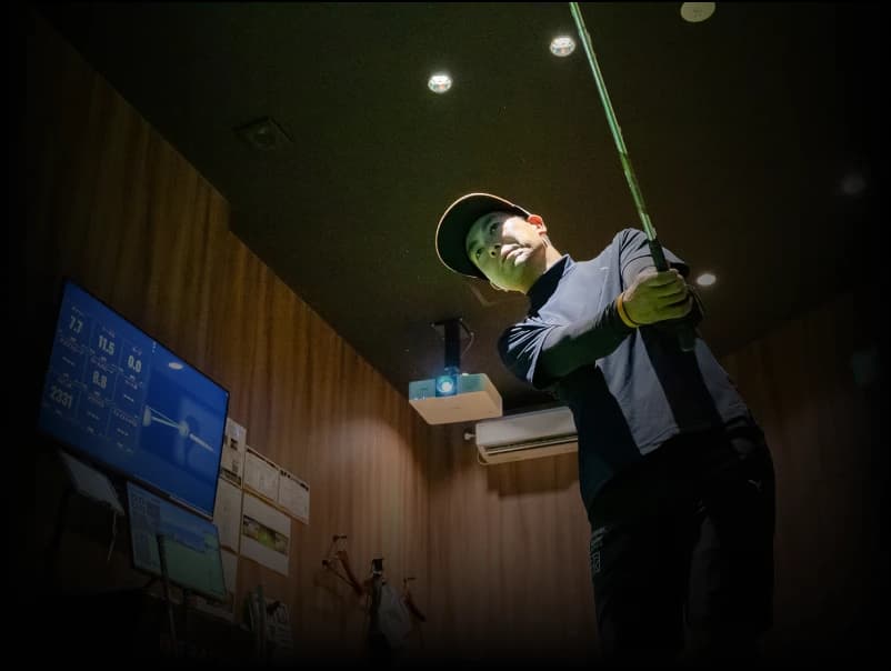 Person in a golf simulator room holding a club, with data screens and a projector visible in the background.