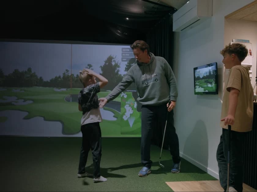 A man with two boys in a golf simulator room, holding a golf club, while a golf course is projected on the screen in the background.